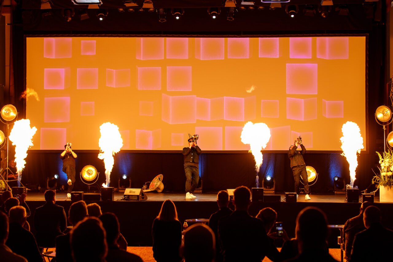 Tre musikere spiller på blåseinstrumenter, mens flammeeffekter lyser opp scenen. Foto.