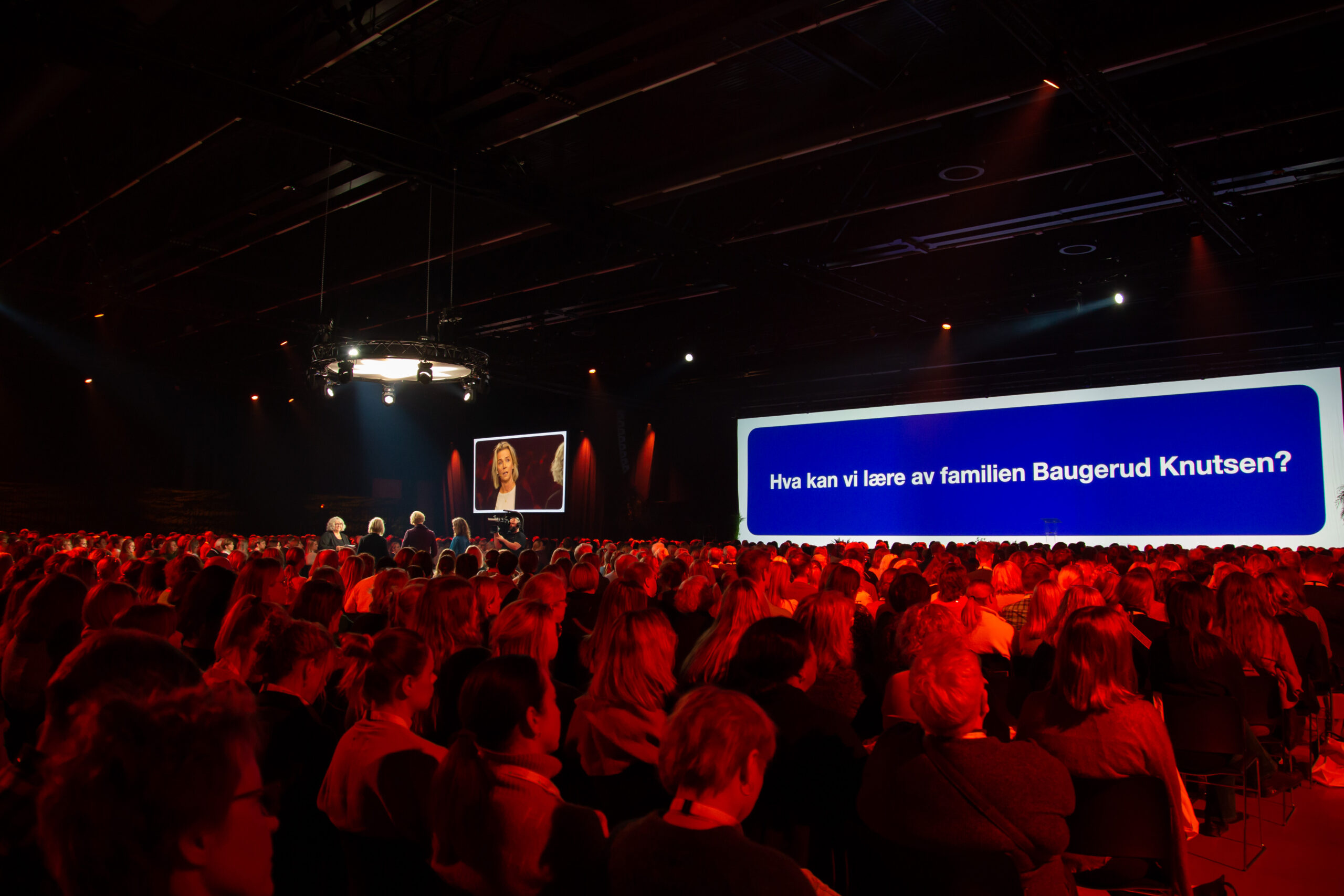 Foredragsholdere står midt blant fullsatt konferansesal. Foto