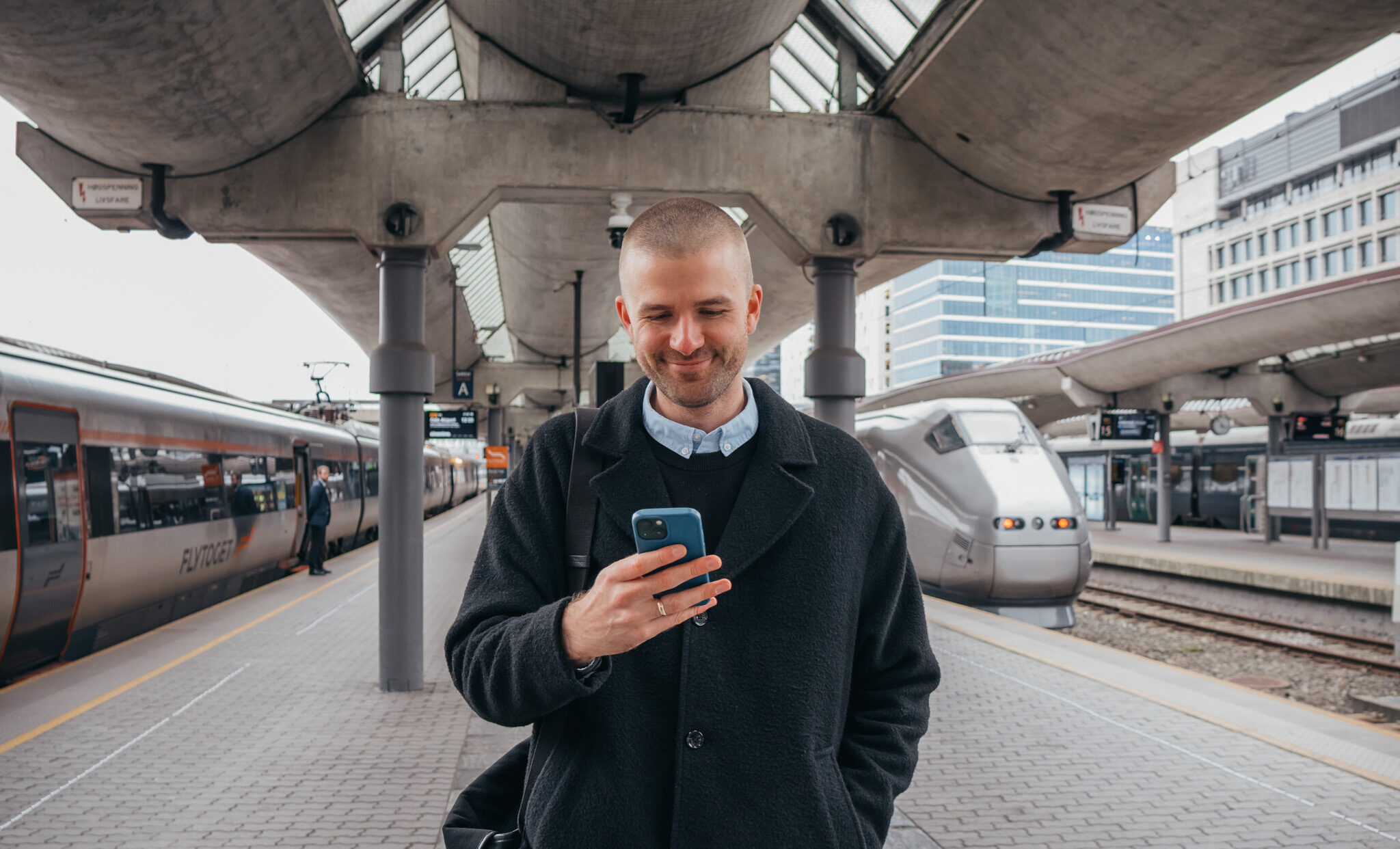 Man på togstasjon ser inn i mobil og smiler. Foto.