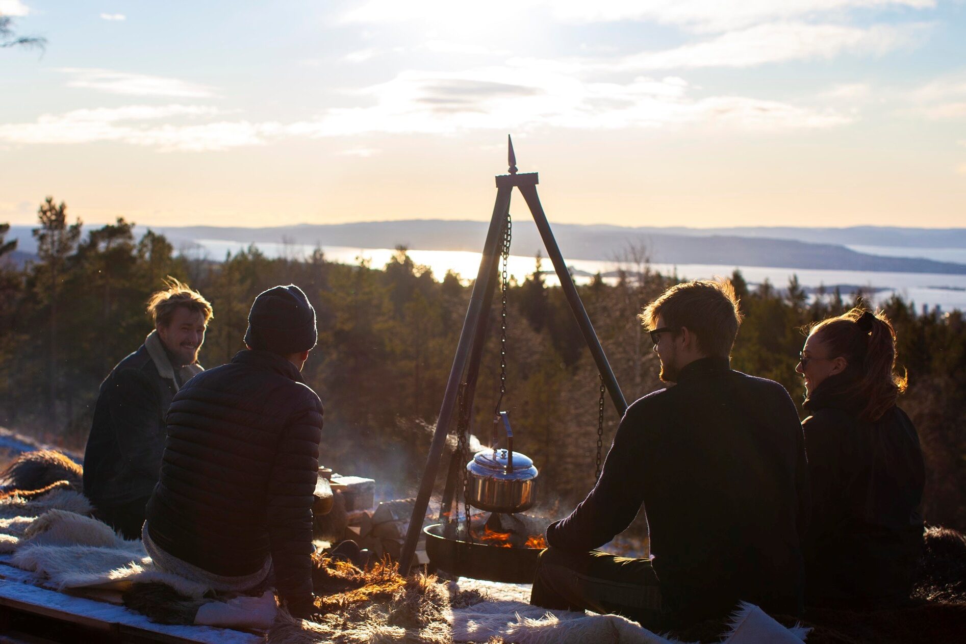 Mennesker sitter rundt bålpanne med utsikt over skog og by. Foto