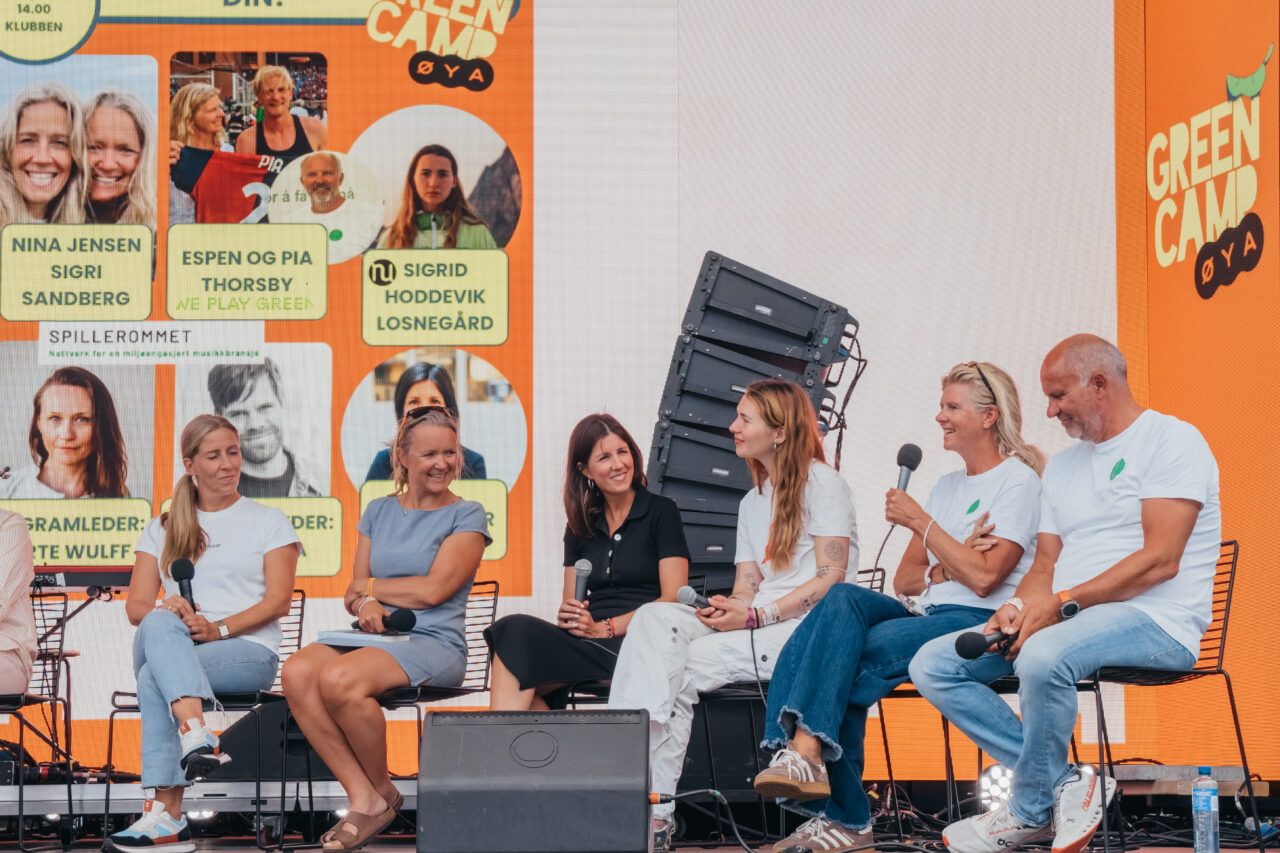 Seks personer sitter på hver sin stol på en scene, vendt mot publikum. Foto.