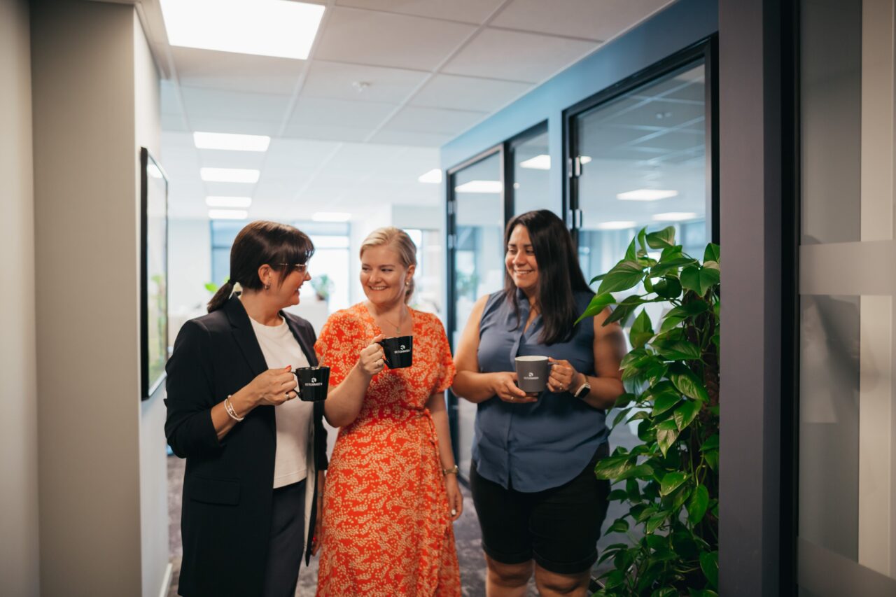 Tre personer står i en kontorgang og holder kaffekopper mens de prater sammen.
