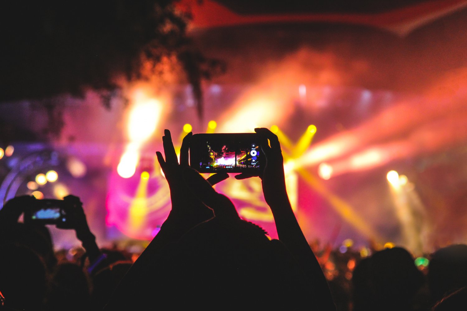 En person holder mobilkamera opp og filmer på en konsert med lyskastere fulle av farger