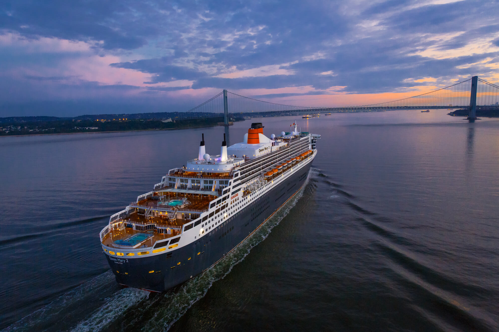 Cruiseskipet Queen Mary 2 i solnedgang på havet, seiler mot en bro i horisonten, på vei ut fra New York. Foto.