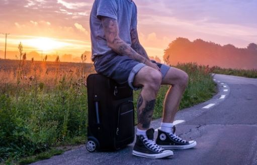 Mann med tatoveringer sitter på en trillekoffert, langs landeveien