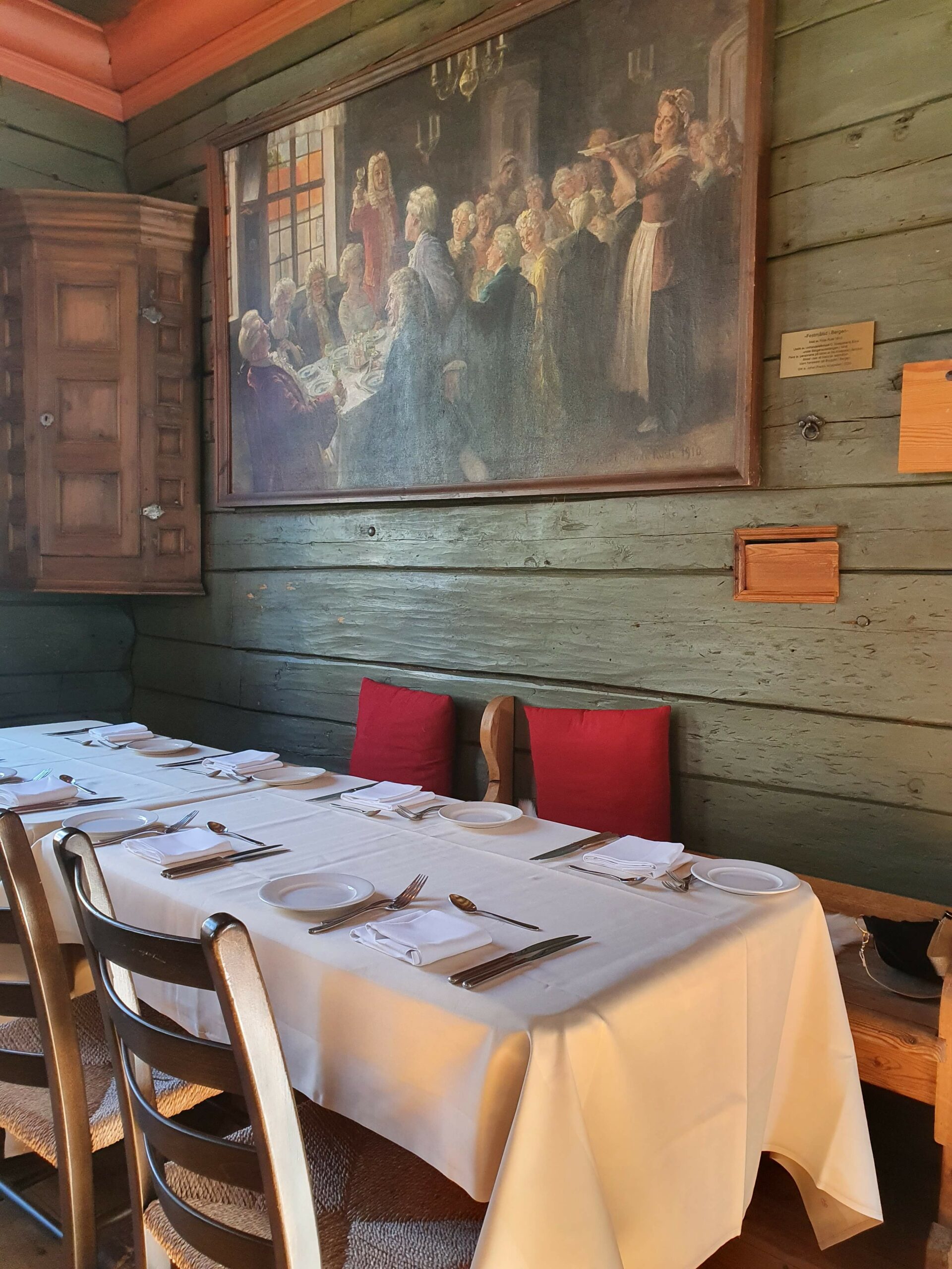 Gammel stue med laftede grønne vegger, pådekket langbord og et gammel maleri i bakgrunnen