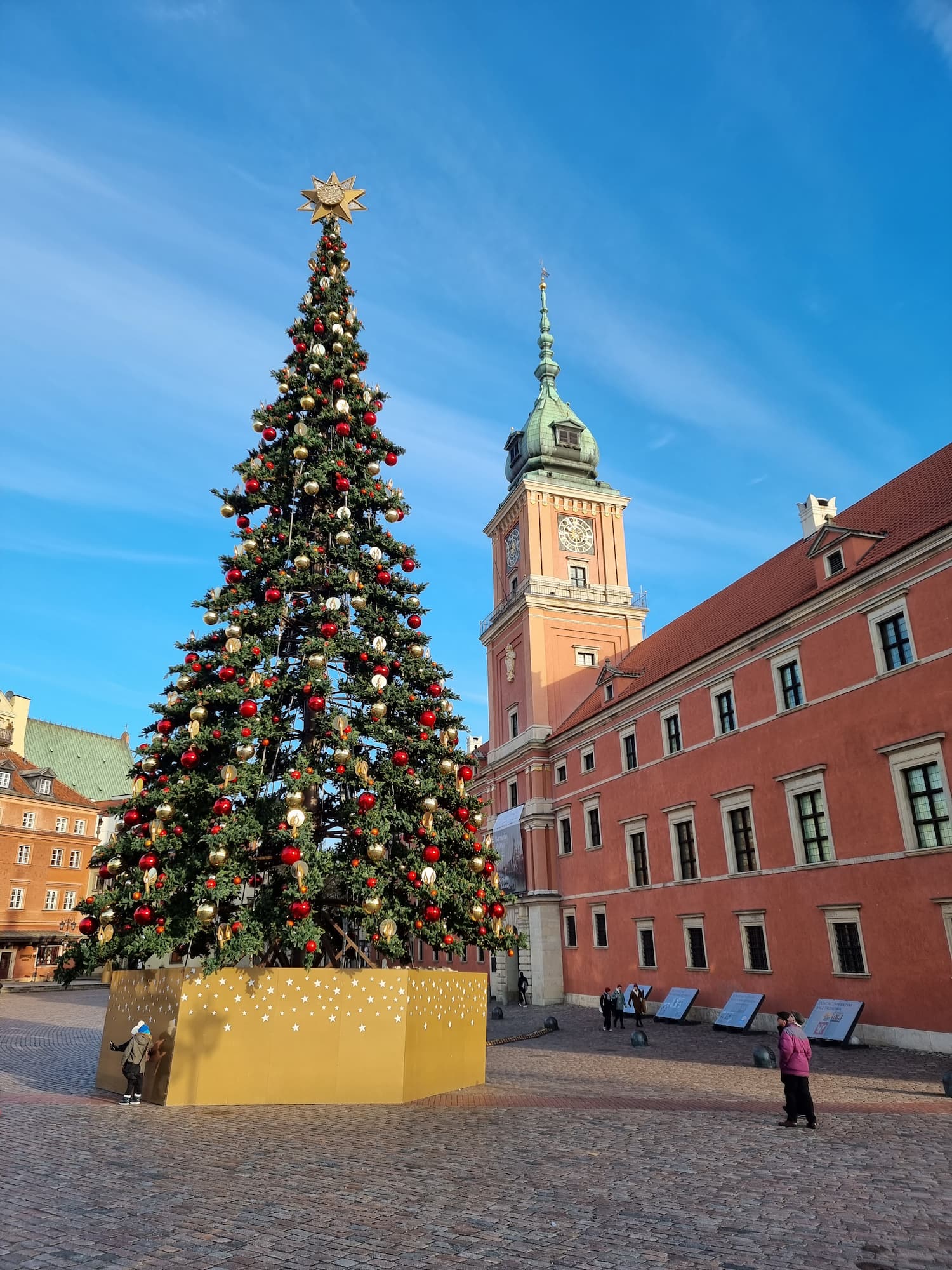 Et stort pyntet juletre foran et ferskenfarget palass med et høyt tårn. Foto.