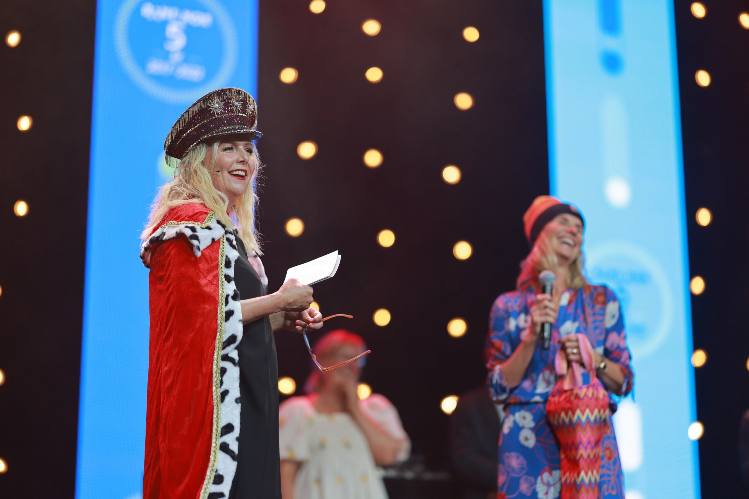 Kvinne kledd i rød kappe og hatt står på en scene. Foto