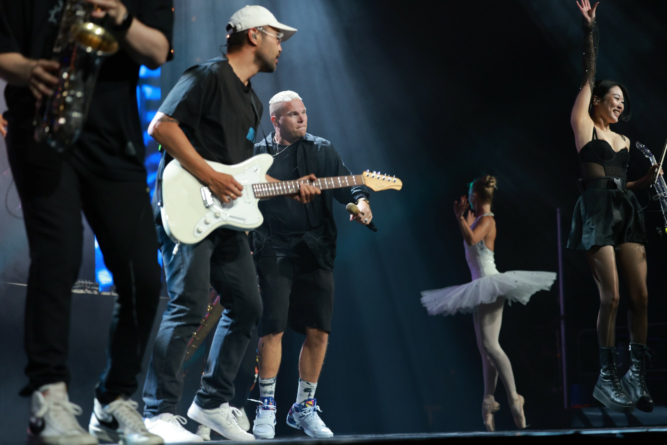 En ballerina i hvitt står og danser blant musikere på en scene. Foto