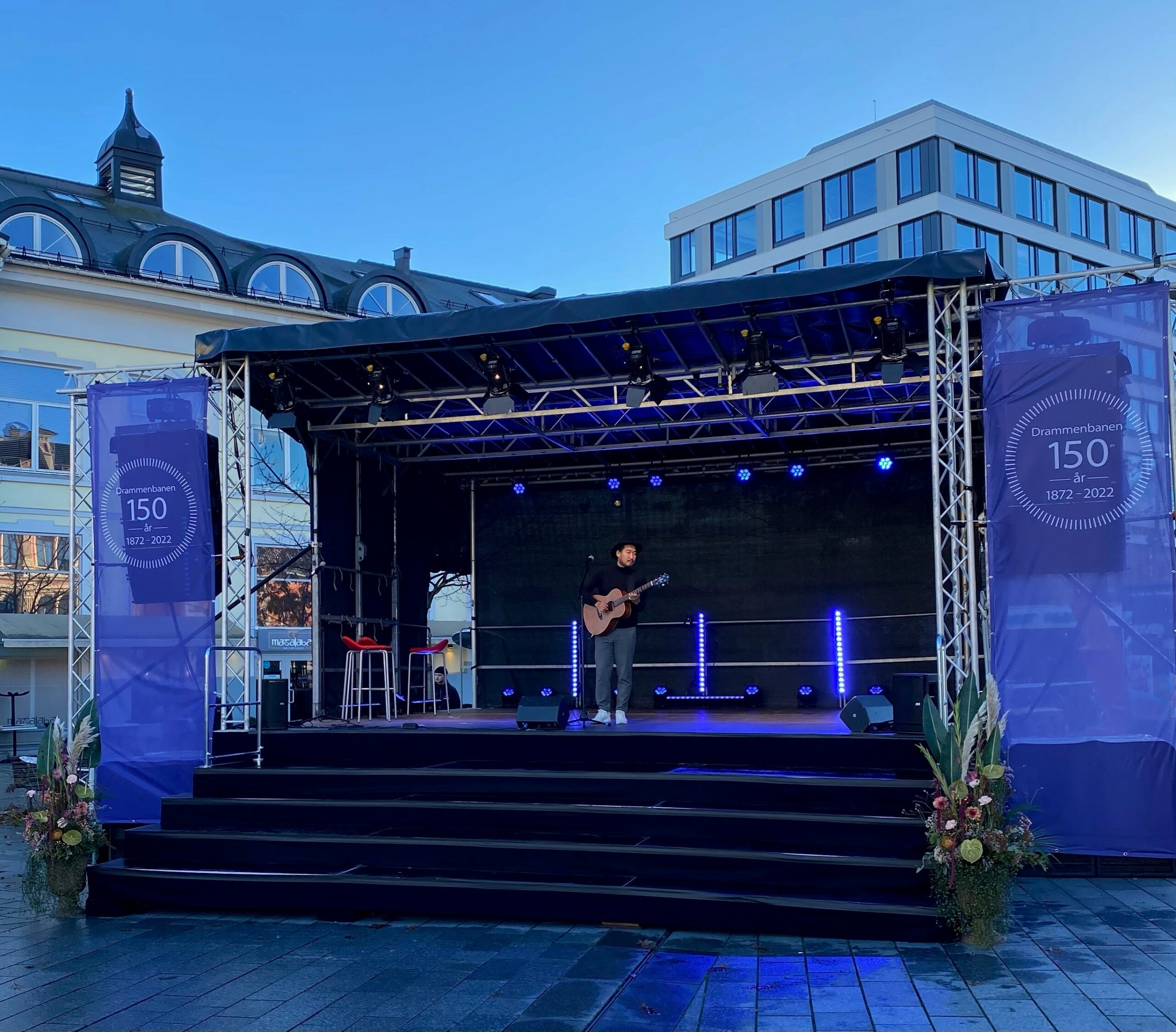 En gitarist står på en scene og spiller på et torg. Foro