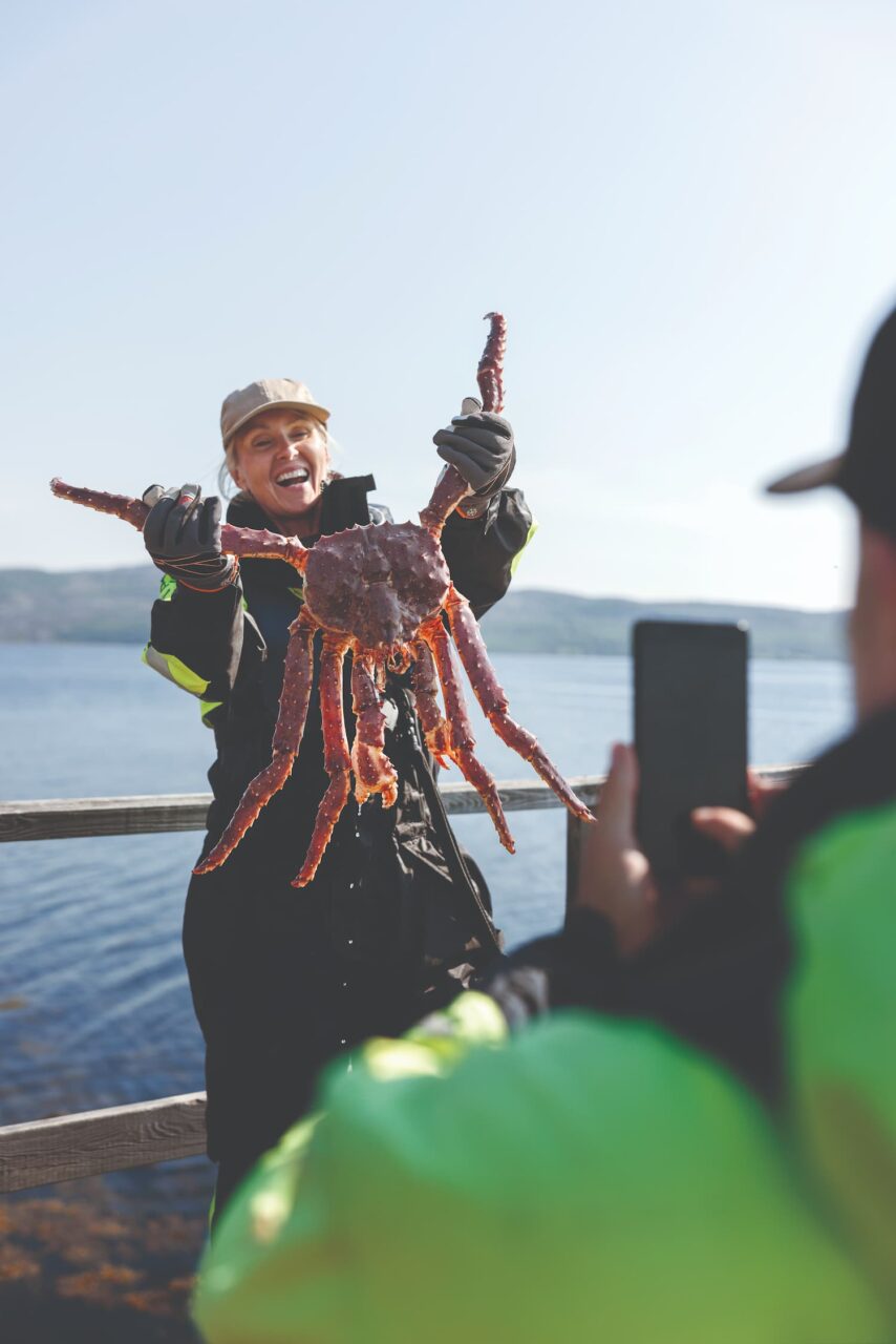 Kvinne holder opp stor kongekrabbe mens hun smiler bredt. Foto