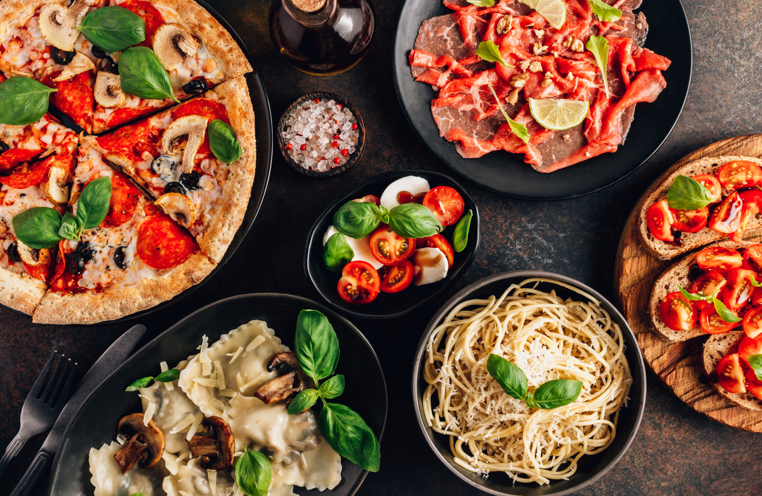 Pizza, pasta og grønnsaker i diverse fat på mørk bordplate. Foto