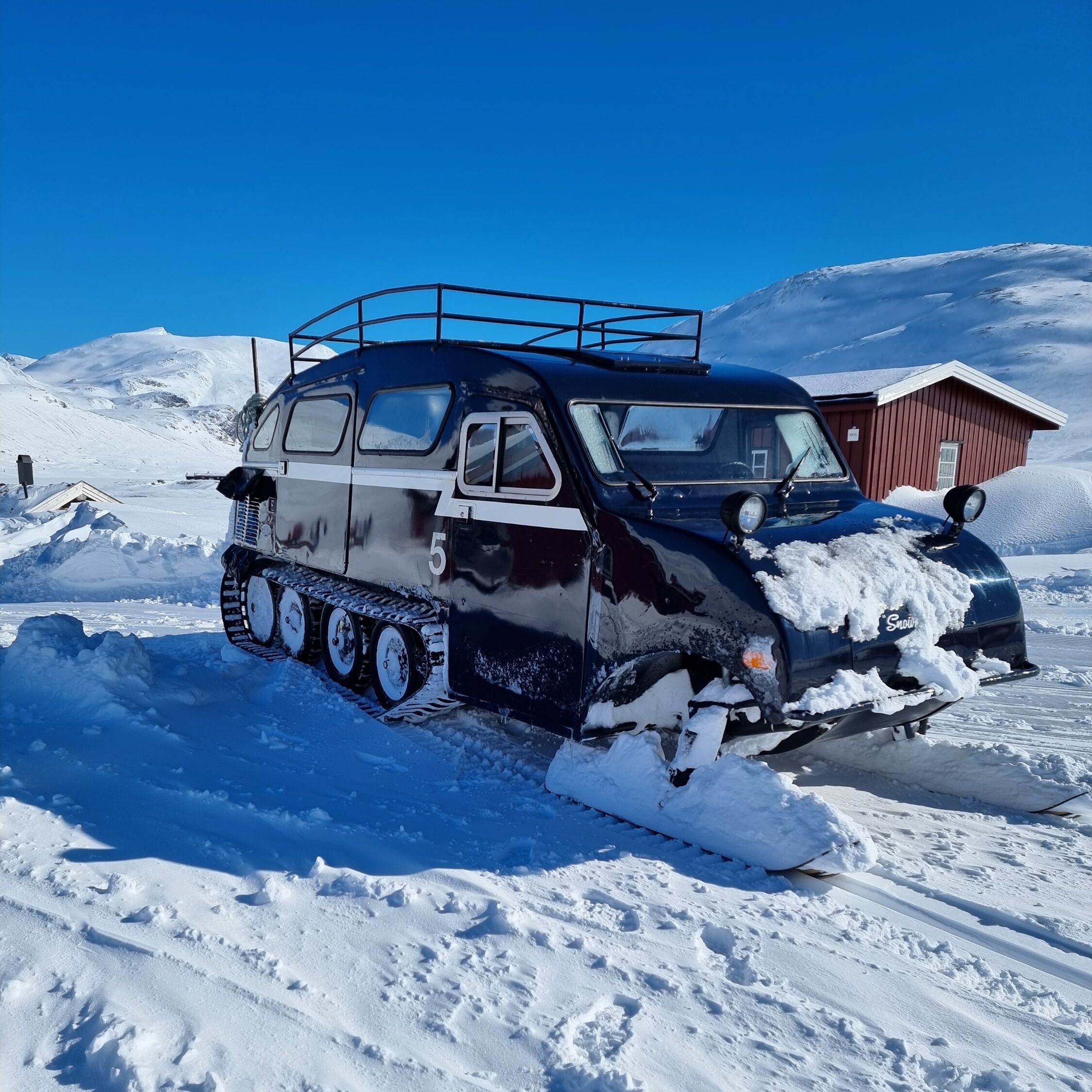 Blå beltevogn omgitt av snø og fjell. Foto
