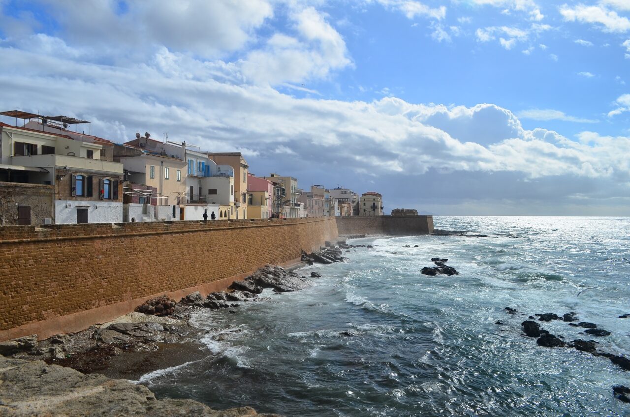 Alghero med fargerike steinhus til venstre, blått havet til høyre. Blå himmel med hvite skyer. Foto energetta - pixabay