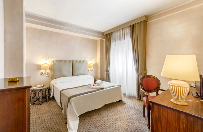 Colonna Palace Mediterraneo - Stor dobbeltseng med hvitt sengeteppe. Utgang til balkong. Skrivebord med rød stol, og hvit lampe. Foto WebBeds