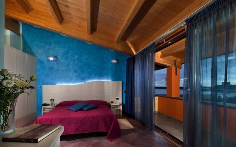 Hotel Al Gabbiano - Dobbeltseng med lilla sengeteppe og blå, små, pynteputer. Glassdør ut til stor balkong. Foto WebBeds