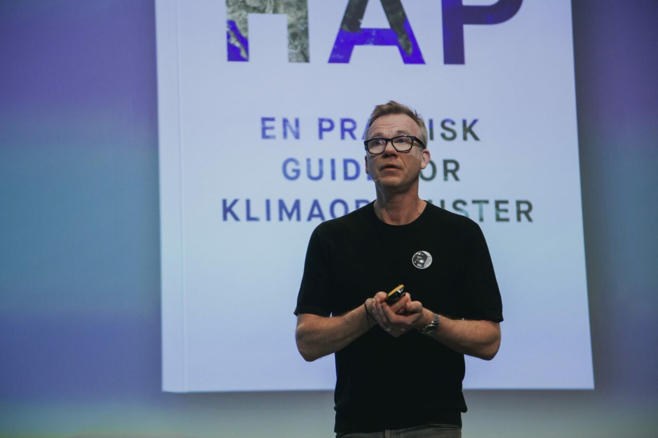 Mann står på scene med hendende foldet og ser engasjert ut mot kamera. Foto