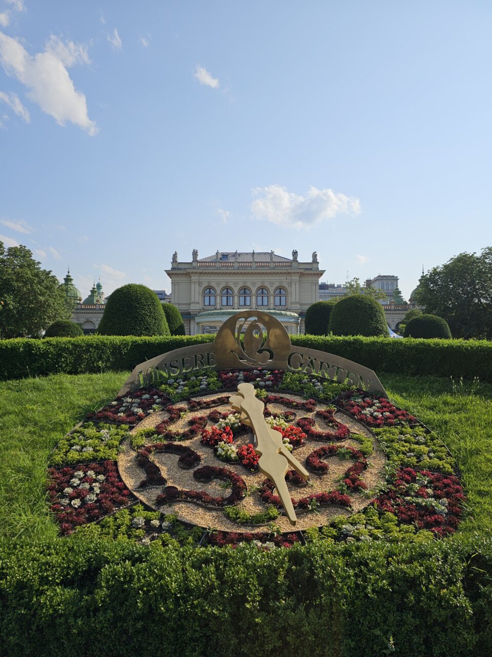 Klokke i et blomsterbed foran en paviljong. Foto